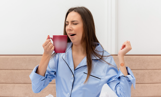 Woman in pyjamas yawning while holding a coffee mug in the morning, wearing a sleep patch on her arm and looking tired after poor sleep.