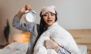Woman in pajamas holding an alarm clock and pillow while sitting on a bed, looking tired after a restless night.