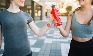 Two women sharing an energy drink after a workout, with one wearing an energy boost patch on her wrist, comparing wearable energy patches with traditional energy drinks for active lifestyles.