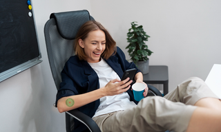 A woman taking a break at work, smiling while using her phone, with a support patch applied to her arm.