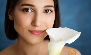 Close-up portrait of a woman holding a white flower with a small round wellness patch visible on her shoulder against a soft blue background.
