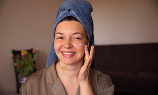 Smiling woman wearing a beauty patch on her neck while applying face cream after a shower, highlighting a skincare routine for healthy, glowing skin.