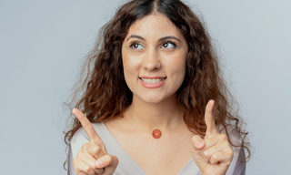 Smiling woman pointing upward with both hands while wearing a wellness patch on her chest against a light background.