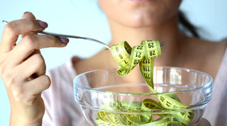 Woman holding a fork lifting a yellow measuring tape from a glass bowl, symbolising weight control and hunger management.