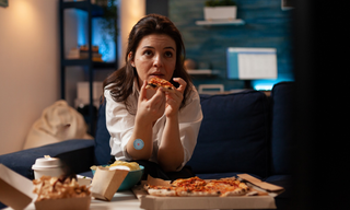 Woman eating pizza late at night while sitting on a sofa, illustrating night-time hunger and late-night snacking.