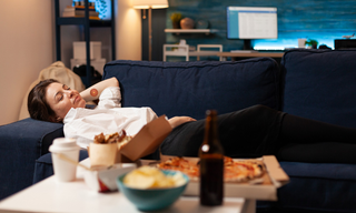 Woman lying on a sofa in a dim living room with a small patch visible on her upper arm; takeaway pizza, snacks, and a drink are on the table in front of her.