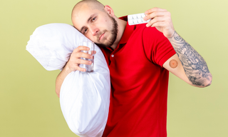 Man holding sleeping pills and a glass of water, wearing a sleep patch on his arm and leaning on a pillow, comparing sleep aids.