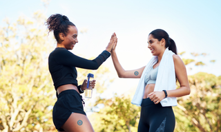 Two women in activewear high-fiving outdoors after a workout, wearing wellness patches and enjoying a healthy, active lifestyle with hydration and fitness support.