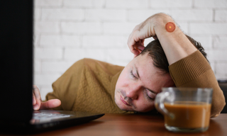 Tired man resting his head on a desk beside a laptop and coffee mug, showing signs of afternoon fatigue while wearing an energy patch.