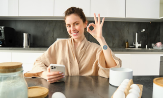 Smiling woman wearing a wellness patch on her wrist while using a smartphone in her kitchen, representing an easy daily wellness routine with wearable health patches for beginners.