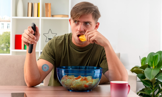 Man eating crisps while watching TV with a wellness patch on his arm, illustrating afternoon snacking habits and cravings.