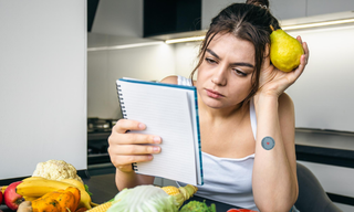 Woman sitting in a kitchen holding a pear and reading a notebook, looking confused about healthy eating choices, with a patch on her arm.