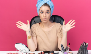 Woman with a towel on her head looking surprised, wearing a skincare patch, sitting at a table with makeup products.