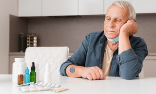 Older man sitting at a table with supplements and wellness products, looking tired while wearing a wellness patch on his arm.