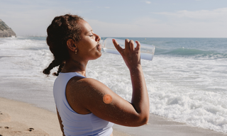 Woman drinking water on a beach while wearing a wellness patch on her upper arm, promoting hydration, healthy living, and outdoor wellness support.