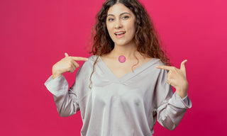 Young woman with curly hair smiling and pointing to a pink wellness patch applied to the front of her neck, standing against a bright pink background and wearing a light grey blouse.