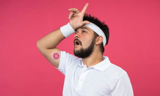 Man in a white polo and headband on a pink background, holding his hand to his forehead in a frustrated “loser” gesture, with a small patch on his upper arm.