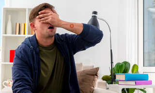Exhausted man covering his face while sitting at home, showing low energy and fatigue while wearing an energy patch.