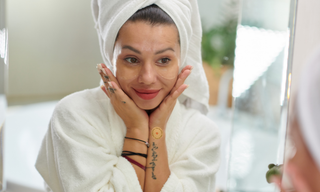 Woman in a white bathrobe and towel looking in the mirror with under-eye patches applied, wearing a wellness patch on her wrist as part of a simple self-care and skincare routine.