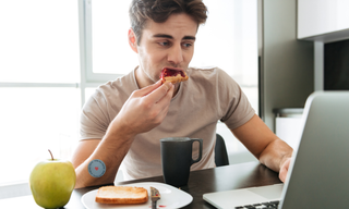 Person eating toast at a desk despite having already eaten, representing feeling hungry after eating and appetite imbalance.